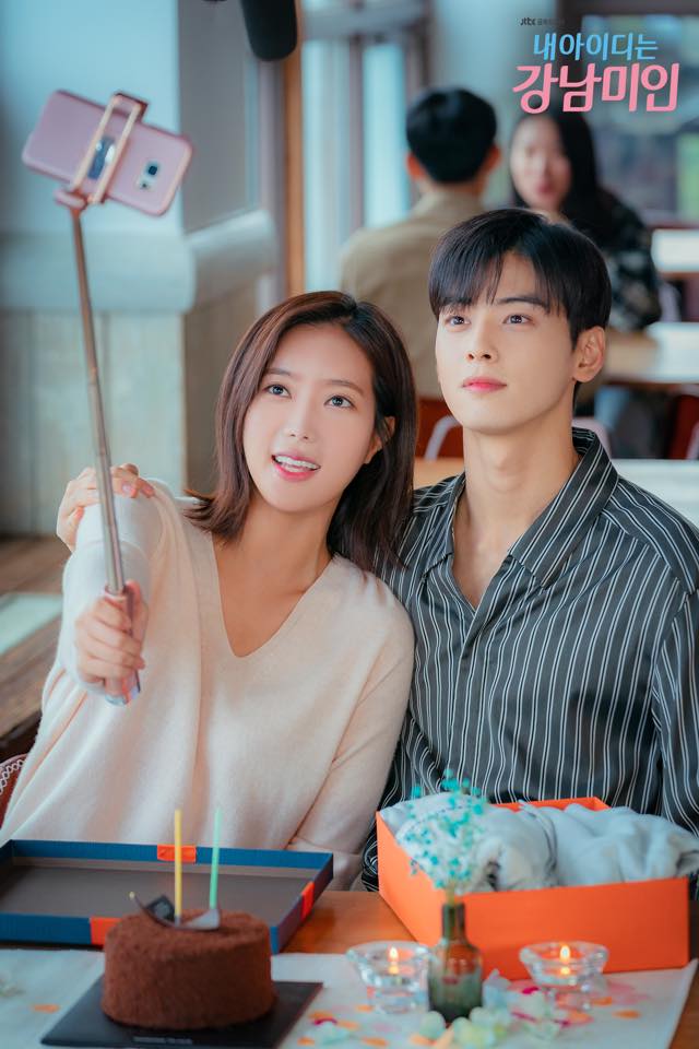 A still from the drama My ID is Gangnam Beauty. Characters Kang Mi-rae and Do Kyung-seok sit closely side by side in a cafe. Mi-rae holds a selfie stick with a pink smartphone to take a photo of them together, while Kyung-seok looks up toward the camera with a subtle expression. In the foreground, a small chocolate birthday cake with candles and a gift box sit on the table.
