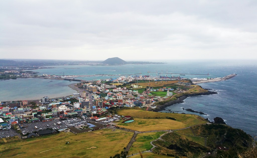 An aerial landscape view on an overcast day on jeju island looking down on a coastal town with Udo island visible on the horizon.