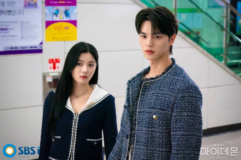 Actor Song Kang and actress Kim Yoo-jung stand side by side in a still from the South Korean drama My Demon. Song Kang stands in the foreground on the right, looking off-camera with a focused expression, wearing a navy blue textured, frayed tweed jacket. Kim Yoo-jung stands slightly behind him on the left with long dark hair, wearing a matching navy jacket with white trim and a white collar, looking in the same direction with a serious, curious expression. Colorful promotional posters hang on the wall in the background.