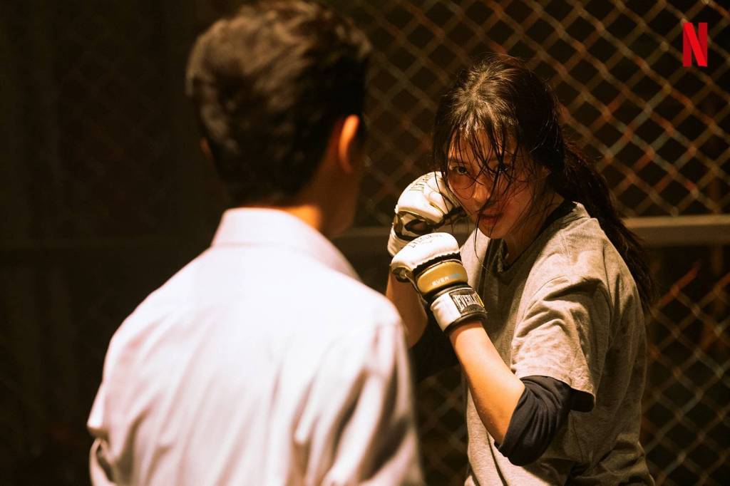 An intense scene from Netflix's "My Name" showing Ji-woo (Han So-hee) wearing white Everlast boxing gloves and a gray t-shirt, staring down her opponent in a dimly lit boxing ring surrounded by chain-link fencing. Her face and hair are drenched in sweat and blood.