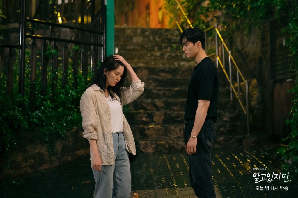 Medium shot of actors Han So-hee and Song Kang standing on a wet street at night with stone steps in the background, from the drama Nevertheless. Han So-hee wears a light-colored button-up over a white shirt and light jeans, running a hand through her wet hair while looking down. Song Kang stands opposite her in a black short-sleeved polo shirt, looking at her intently.