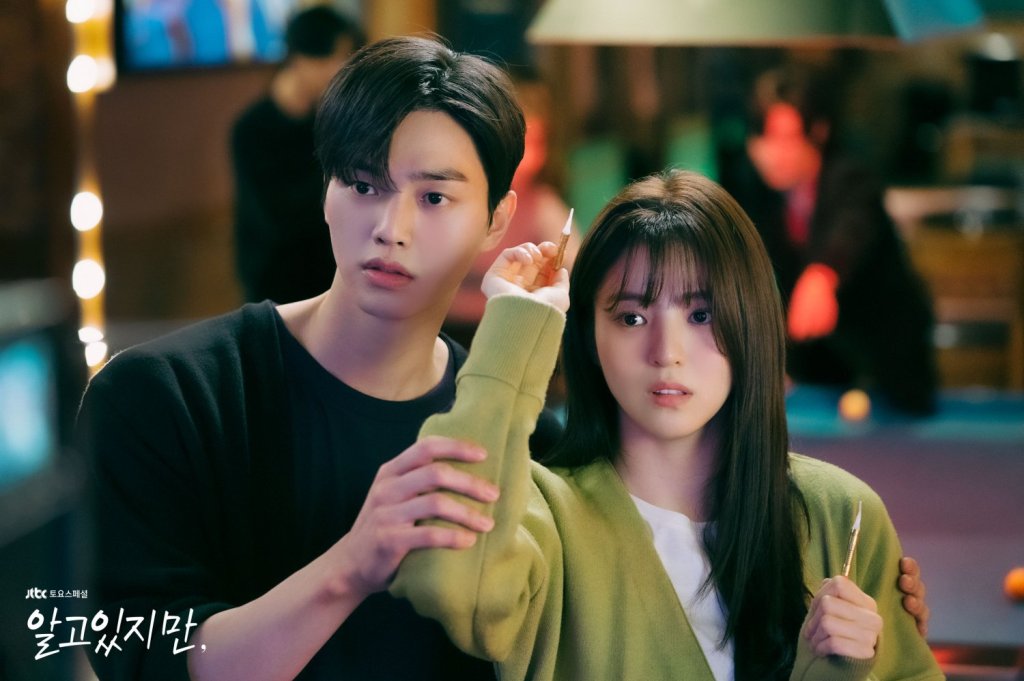 Close-up shot of actors Song Kang and Han So-hee from the Korean drama Nevertheless. Han So-hee stands in the foreground wearing a light green cardigan, holding a dart and looking ahead with an anxious expression. Song Kang stands closely behind her in a black top, gently reaching around her to guide her hand.